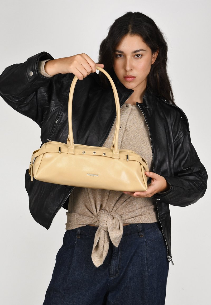 Bolso de cuero beige con asas dobles, forma elegante y cierre de cremallera, sostenido por una persona que lleva una chaqueta de cuero negra y una blusa beige.