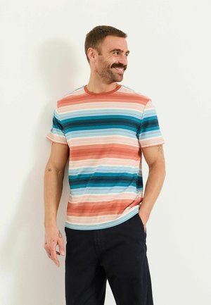 Mann mit Schnurrbart, der ein kurzärmeliges gestreiftes Hemd in Blau- und Orangetönen trägt, schwarze Hose, lächelt und schaut zur Seite.