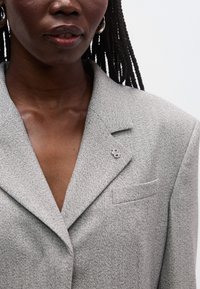 Ανοιχτό γκρι σακάκι με υφή υφάσματος, με γιακά notch, διακριτική τσέπη στο στήθος και διακοσμητική ασημένια καρφίτσα.
