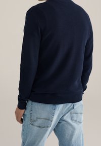 Man i en mörkblå texturerad tröja och ljusblå jeans, sedd bakifrån mot en enkel bakgrund.