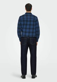Homme portant une chemise à manches longues à carreaux bleus et noirs, un jean bleu marine et des chaussures noires, se tenant de dos devant un fond uni.
