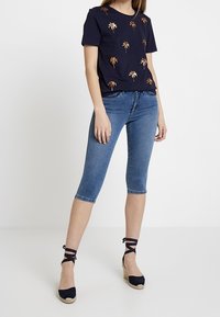 Námořnické tričko s vyšitým zlatým palmovým stromem, spojené se světle modrými capri džíny a námořnickými espadrilkami na klínu s kotníkovými pásky.