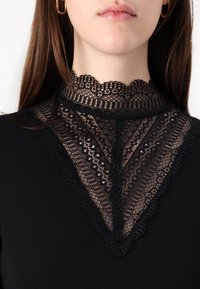 Svart topp med hög spetskrage som har intrikata mönster och ribbad textur. Spetsen ligger över en solid svart tygbas.