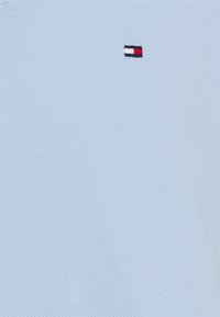 Vaaleansininen polo-paita, joka on valmistettu teksturoidusta kankaasta ja jossa on pieni brodeerattu logo punaisena, valkoisena ja mustana vasemmassa rinnassa.