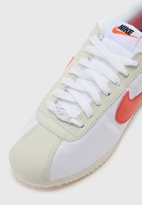 Nike sportbačiai su balta tekstiline viršutine dalimi, pilkais zomšo akcentais, oranžiniu logotipu ir plokščiomis baltomis raištelėmis. Gumos padas su tekstūruota danga.