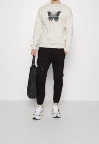 Krämfärgad sweatshirt med svart fjärilstryck, svarta cargo-byxor med dragkedjefickor och vita sneakers med svarta detaljer, håller en svart tote-väska.