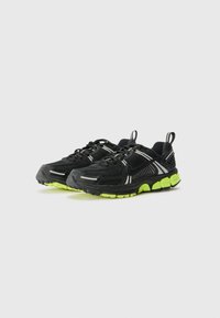 Baskets de sport noires avec un upper en mesh, des accents argentés et une semelle verte. Les caractéristiques incluent un système de laçage et une languette à l'arrière.