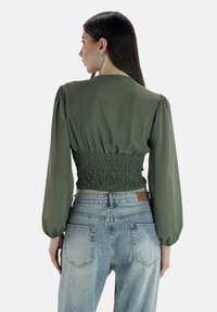 Olivgrüne Bluse mit Puffärmeln und geraffter Taille. Das Material wirkt glatt und halbtransparent, kombiniert mit hellblauen Jeans.