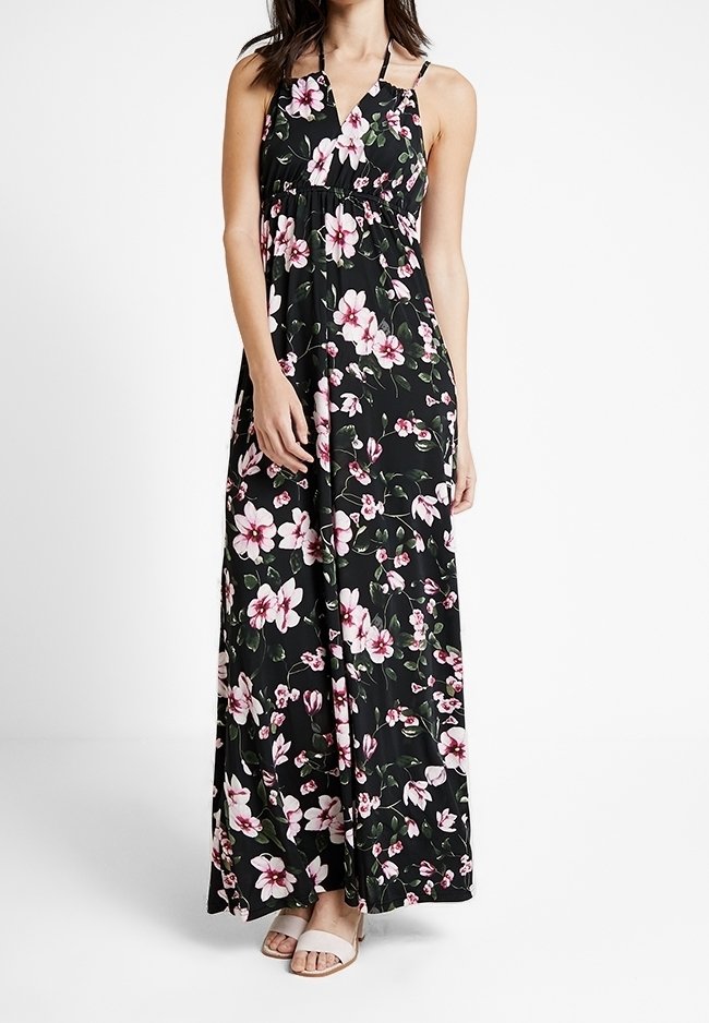 Femme portant une robe maxi noire à fleurs roses et blanches, bretelles spaghetti et sandales ouvertes de couleur claire.