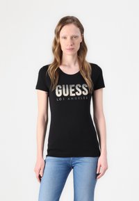 Melnā pieguļošā T-kreklā ar īsām piedurknēm; uzdrukāti vārdi "GUESS" un "LOS ANGELES" metaliskā fontā. Modelis ģērbies zilās džinsa biksēs. Vienkāršs fons.