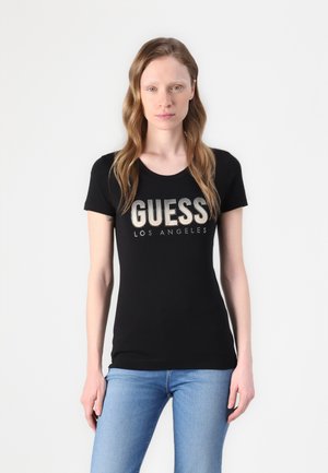 Črna oprijeta majica s kratkimi rokavi; ima napise "GUESS" in "LOS ANGELES" v kovinskih črkah. Model nosi modre kavbojke. Enostavna ozadje.