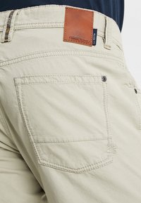Pantalones cargo beige con un bolsillo trasero con detalle de costura, etiqueta de parche de cuero y cierre de botón. Material de algodón suave y duradero.