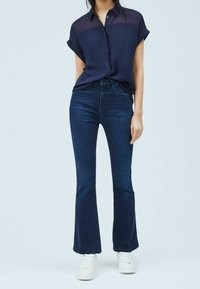 Navy short-sleeve blouse with sheer upper panels, paired with slim-fit dark denim flared jeans and white sneakers on a light background.