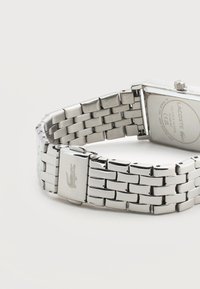 Bracelet de montre en métal argenté avec fermoir arborant le logo du crocodile Lacoste et boîtier rectangulaire au dos de la montre affichant la marque et les détails.
