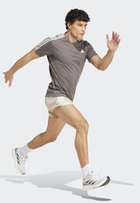 Camiseta deportiva gris con rayas blancas, pantalones cortos beige y zapatillas de correr blancas. El sujeto está en movimiento, con un enfoque en la forma y el movimiento dinámicos.