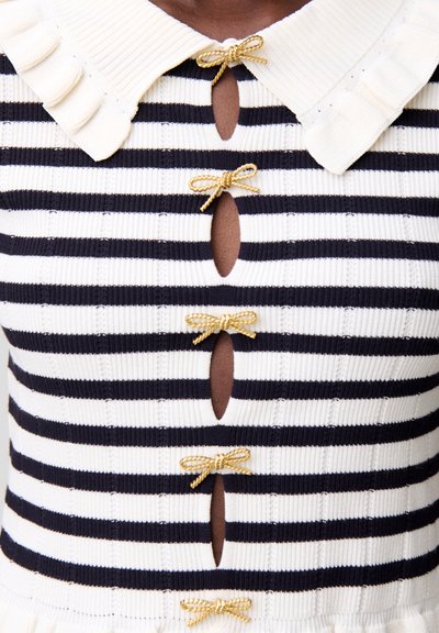 Top de punto a rayas blancas y negras con cuello blanco con borde ondulado y botones frontales en forma de lazo dorado.
