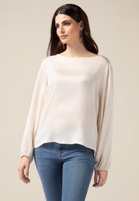 Cremefarvet bluse med lange, bløde ærmer, en bådhals og en let buet kant. Båret med blå jeans, glat tekstur.