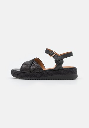 Sort åben tå sandal med vævede stropper, ankelspænde og platformsål med flettet detalje mod en ensfarvet lys baggrund.