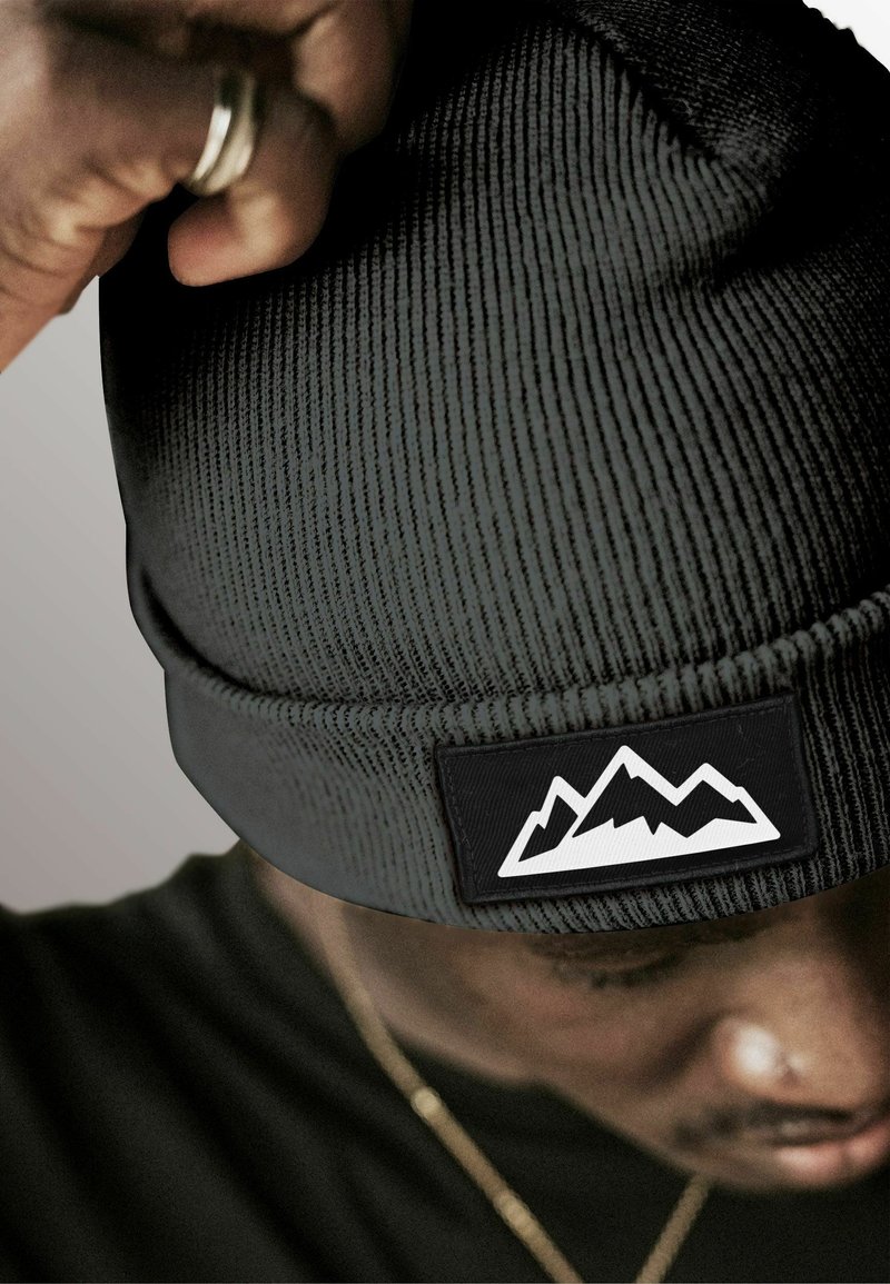 Black ribbed knit beanie featuring a black patch with white mountain graphic. Close-up view shows texture and details of the material.