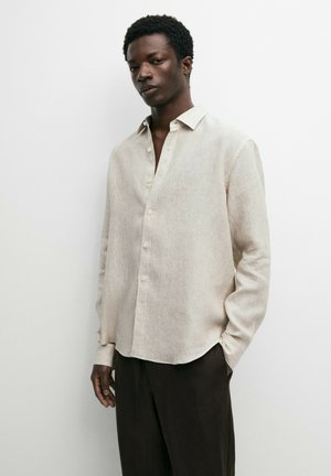 Jeune homme portant une chemise beige à manches longues boutonnée et un pantalon sombre, debout devant un fond blanc uni.