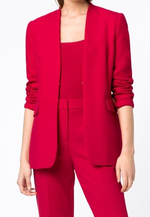 Femme portant un blazer rouge vif avec des manches froncées, un pantalon taille haute assorti et un haut rouge à encolure carrée.