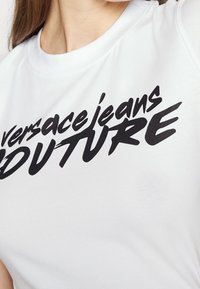 Vit bomull T-shirt med framträdande svart text som läser "versacejeans COUTURE," med en slät yta och rundad halsringning.