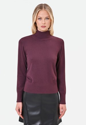 Donna con capelli biondi che indossa un maglione a collo alto viola e una gonna in pelle nera, in piedi davanti a uno sfondo bianco semplice.
