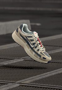 Bež i mornarsko plava Nike tenisica za trčanje s bijelim logotipom i crvenim detaljem na vezicama, suspendirana iznad metalne rešetke na uličnoj gradskoj pozadini.