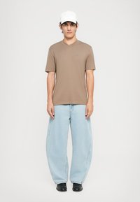 T-shirt marron à manches courtes en V, jean en denim bleu surdimensionné et chaussures noires, complétés par une casquette de baseball blanche.
