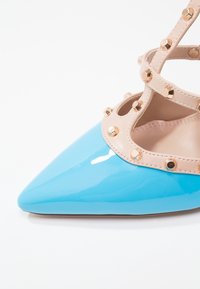 Chaussure pointue en cuir verni bleu avec des sangles beiges cloutées formant un design en T sur le dessus du pied.