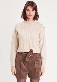 Beige kabelstrikket sweater med høj hals og guld-knapdetaljer på skulderen, parret med brune fløjlskurte shorts med en bundet talje.