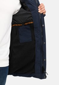 Chaqueta acolchada de color navy con interior de forro polar negro, cuenta con un bolsillo en el pecho y herrajes plateados; cremalleras a lo largo del delantero y los puños.