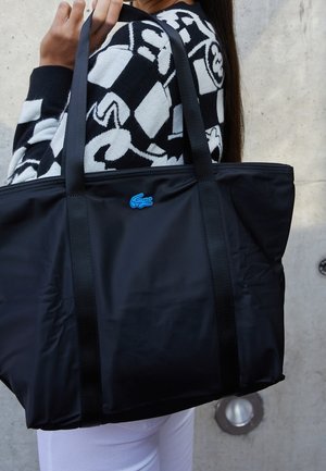 Černá taška z nylonu s dvěma černými uchy, zdobená modrým logem Lacoste. Hladká textura, minimalistický design a horní zapínání na zip.