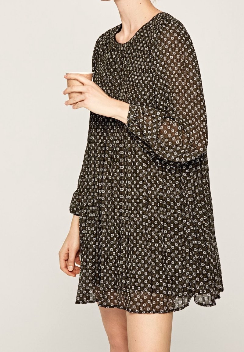Femme portant une robe noire à manches longues avec un motif floral blanc, tenant une petite tasse dans sa main droite sur un fond uni.