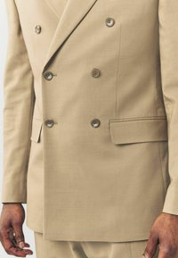 Giacca da uomo doppiopetto beige con due tasche frontali, cinque bottoni e linee pulite. Realizzata in un tessuto strutturato, vestibilità su misura, cuciture sottili.