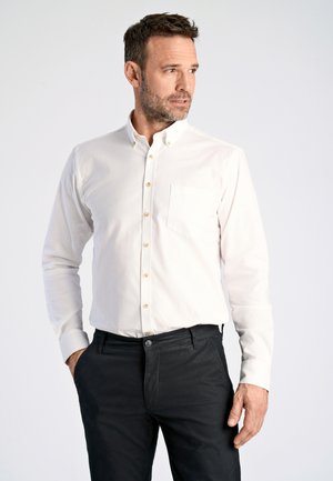Camicia bianca a maniche lunghe con colletto abbottonato e taschino piccolo, caratterizzata da bottoni beige e una texture liscia, abbinata a pantaloni scuri.