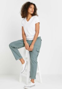 Witte v-hals t-shirt gecombineerd met groene cargobroek, met omgeslagen mouwen en zijzakken. Model draagt witte sneakers. Achtergrond is eenvoudig.