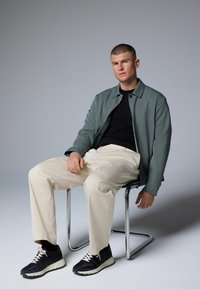 Chaqueta verde abotonada, camiseta negra, pantalones crema y zapatillas negras con detalles en blanco. Modelo sentado en una silla de cromo frente a un fondo gris.