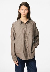 Srajca z gumbi v rjavi barvi z navpičnimi črtami, z enim žepom na prsih, sproščen kroj in dolgimi rokavi. Tkanina se zdi gladka in lahka.