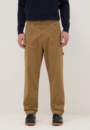 Homme portant un pantalon beige droit et des chaussures noires à lacets, debout devant un fond clair uni.