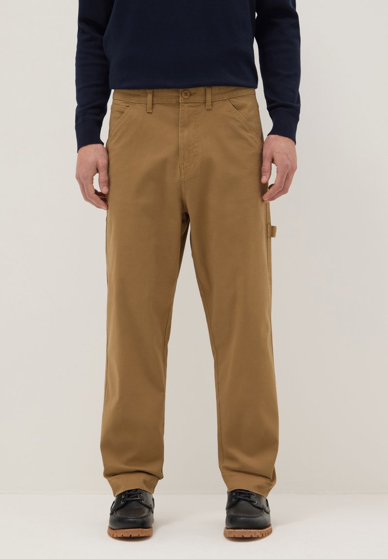 Homme portant un pantalon beige droit et des chaussures noires à lacets, debout devant un fond clair uni.