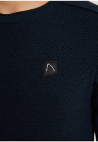 Granatowy sweter wełniany z teksturowanym wzorem i okrągłym dekoltem. Posiada małą czarną łatkę z białym wzorem na piersi.