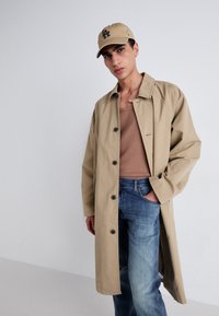 Abrigo trench beige con un diseño de botones en la parte delantera, combinado con una camiseta sin mangas marrón claro y jeans azules. El modelo lleva una gorra beige con detalles en negro.