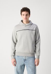 Grå bomullhette med snor i hetten, med svart logo-tekst og en horisontal aksentstripe. Kombinert med lyseblå jeans.
