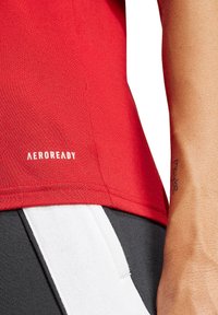 Maglietta da corsa rossa con tessuto testurizzato; presenta il logo bianco "AEROREADY" vicino all'orlo. Include elementi neri e bianchi dei pantaloni vicini.