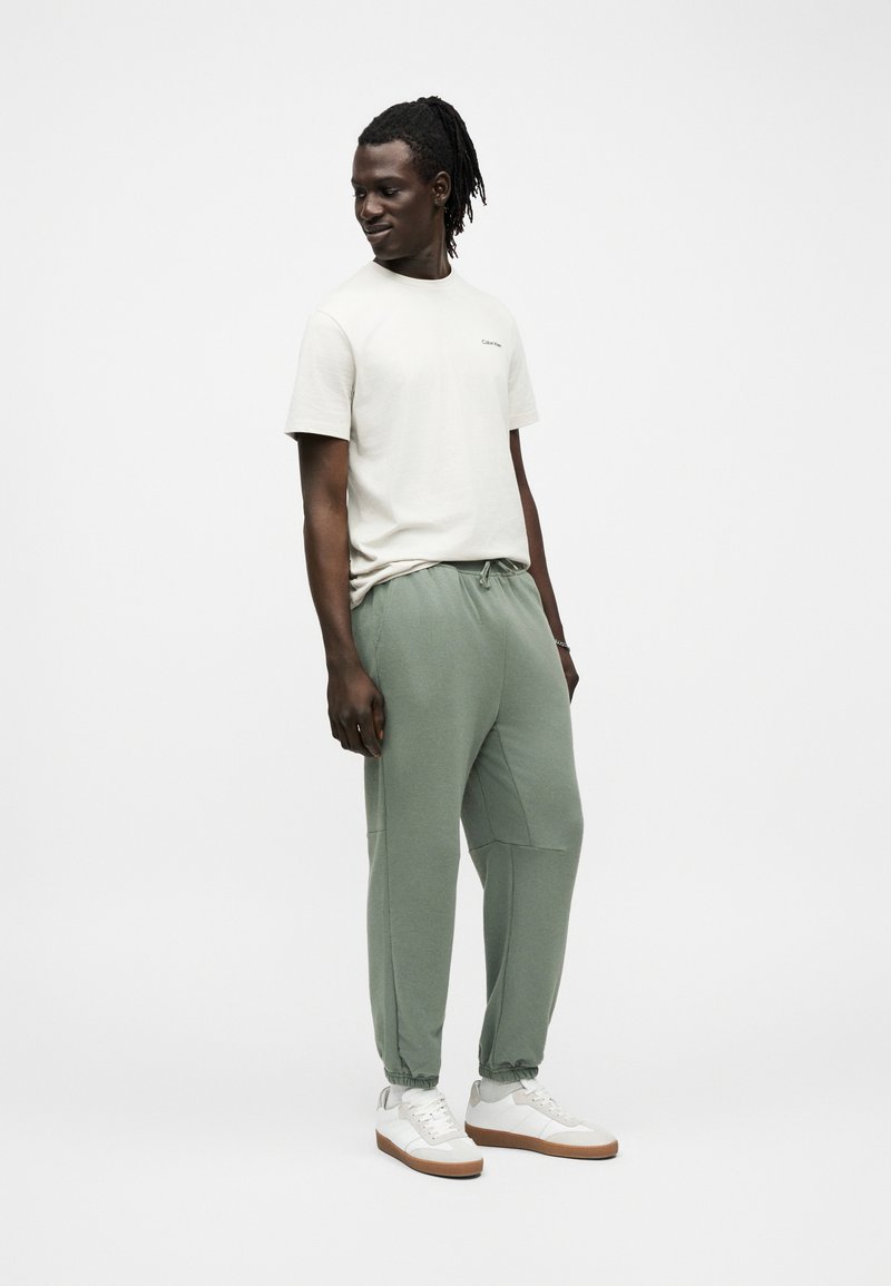 Homme avec des dreadlocks portant un t-shirt blanc, un pantalon de jogging vert sauge et des baskets blanches sur un fond blanc uni.