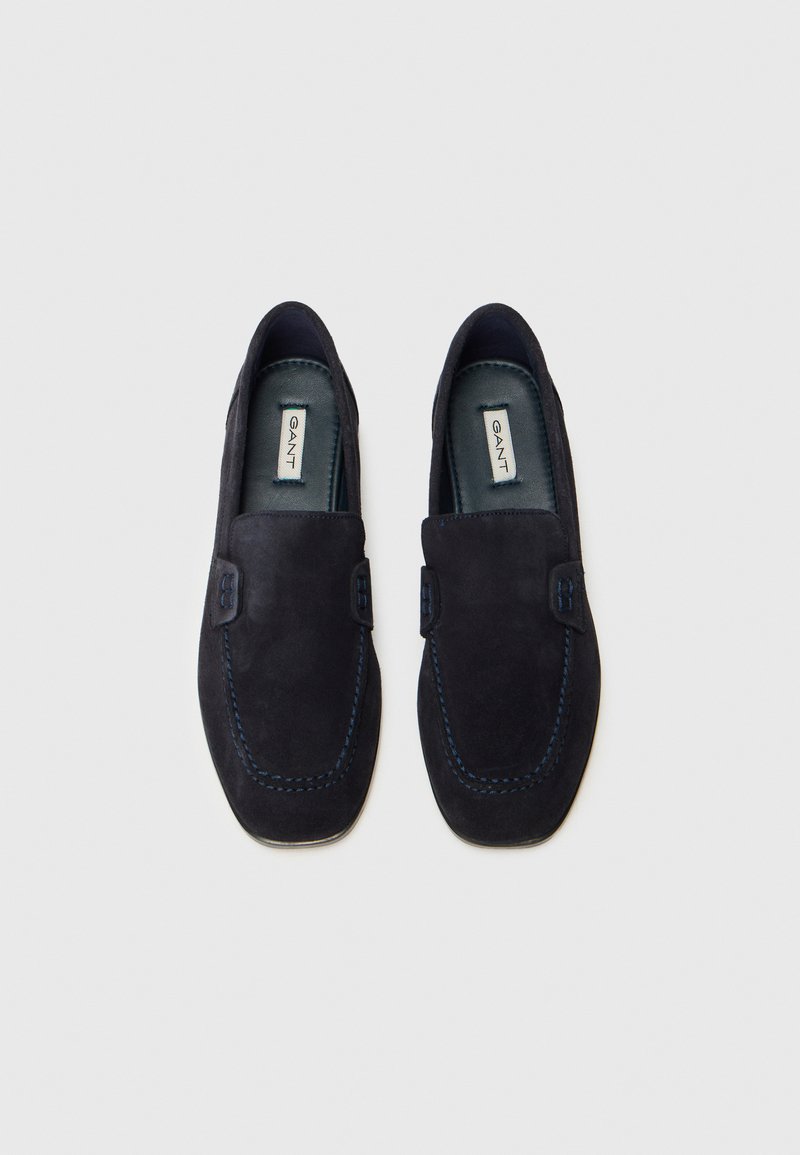 Un paio di mocassini slip-on in suede di un blu navy scuro, con cuciture visibili e etichette GANT sulle sole interne, mostrati dall'alto su uno sfondo bianco.