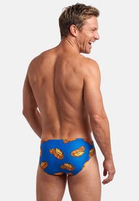 Hymyilevä mies kääntyneenä pois päin, jolla on siniset uimahousut joissa on piirrettyjä tiikerinpäitä, seisoo yksinkertaisen valkoisen taustan edessä.