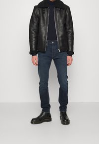 Giacca in shearling nera con chiusura a zip, jeans blu scuro e stivaletti neri. La giacca presenta un collo di pelliccia e una finitura testurizzata.