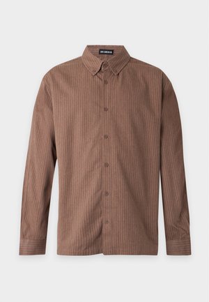 Homme debout sur un fond uni portant une chemise marron à manches longues, un pantalon gris à jambes larges et des baskets noires.